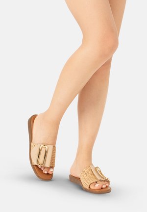 Piernas con sandalias tipo slide beige tejidas con una gran hebilla dorada en una suela marrón claro sobre un fondo neutro.