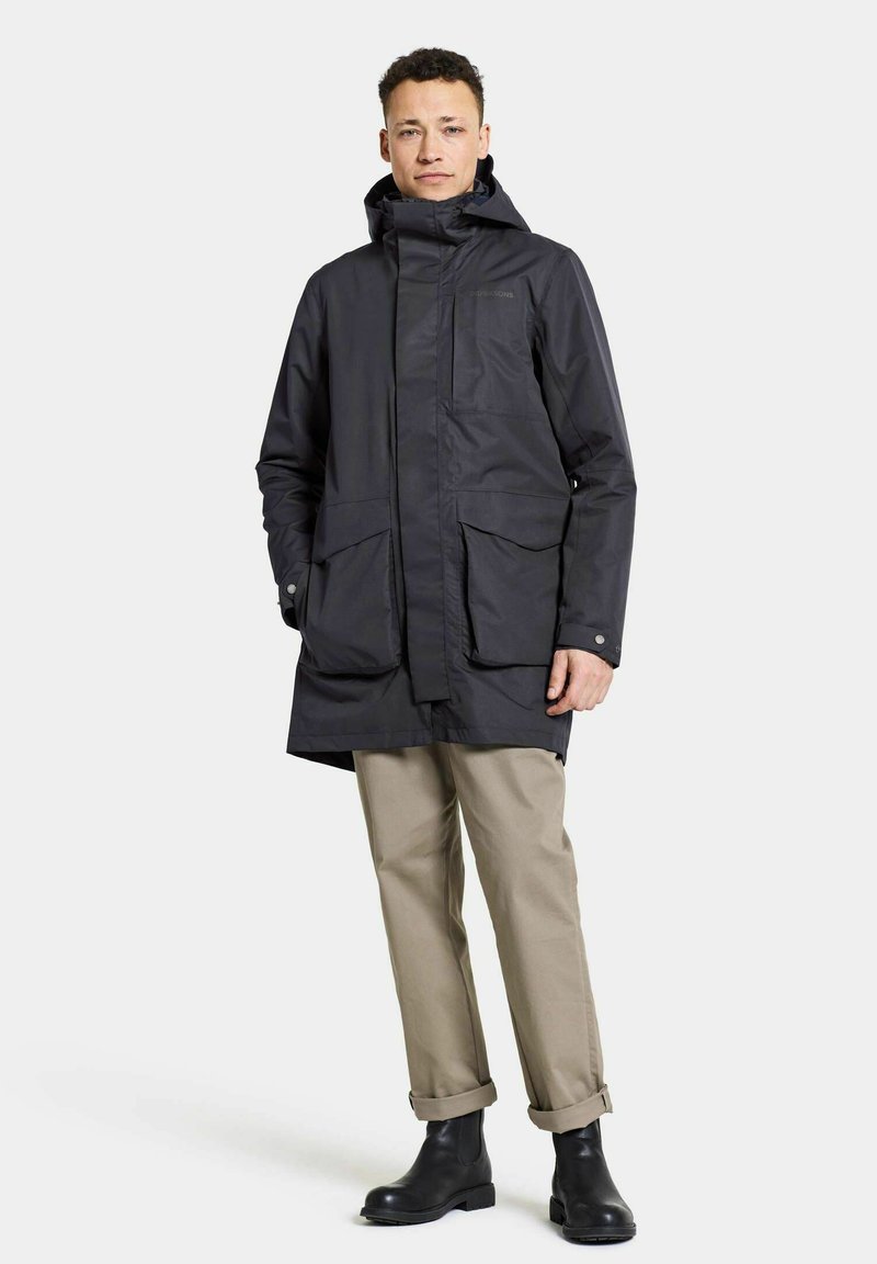Schwarzer, wasserdichter Parka mit Reißverschluss und Druckknopfverschluss, zwei großen Fronttaschen und Kapuze. Getragen mit beigen Hosen und Stiefeln.