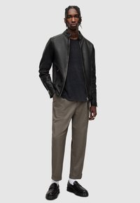 Schwarze Lederjacke mit Stehkragen, kombiniert mit einem dunklen Hemd und hellbraunen karierten Hosen. Schwarze Slipper und weiße Socken.