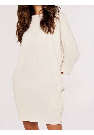 Femme portant une robe ample en maille côtelée crème à manches longues avec poches, debout devant un fond clair uni.