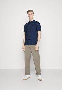 Camicia di lino blu navy a maniche corte, design con bottoni frontali, abbinata a pantaloni chinos beige e sneaker bianche con suola in gomma, in piedi su uno sfondo semplice.