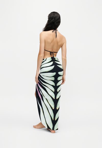 Femme pieds nus portant un haut de bikini noir à col halter et un long sarong noir et blanc à motifs noué à la taille, debout, dos face vers l'avant.
