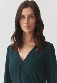 Robe wrap verte avec décolleté en V, manches longues et une texture subtile. Le tissu semble léger, lisse avec un design simple et élégant.