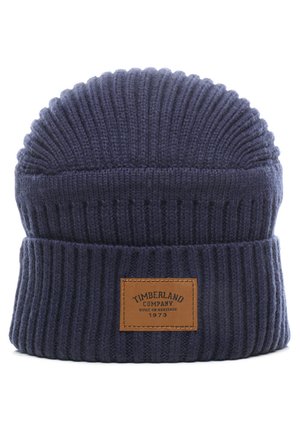 Berretto lavorato a maglia blu scuro con una texture a coste, caratterizzato da una patch in pelle con la scritta "Timberland Company" e un anno, e un design con la parte superiore arrotondata.
