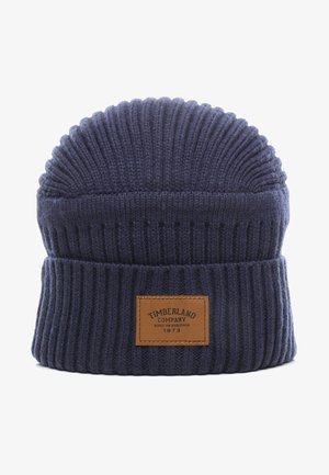 Berretto lavorato a maglia blu scuro con una texture a coste, caratterizzato da una patch in pelle con la scritta "Timberland Company" e un anno, e un design con la parte superiore arrotondata.