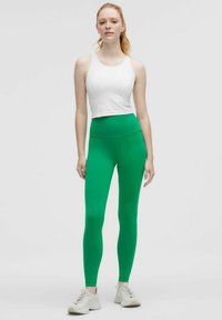 Gröna leggings i stretchigt material, hög midja, som matchas med en vit ärmlös crop top. Modellen har på sig ljusa träningsskor.