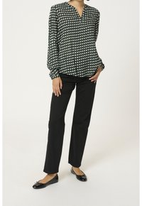 Langärmlige, grüne Bluse mit weißem und pinkem geometrischem Muster, V-Ausschnitt. Kombiniert mit schwarzen geraden Hosen und schwarzen flachen Schuhen.