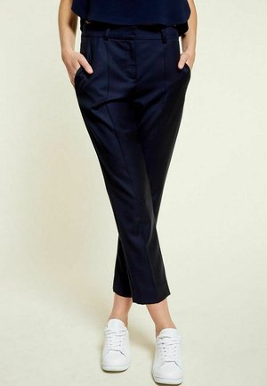 Pantalons taillés navy avec une coupe slim, dotés de poches latérales et d'un ourlet court, associés à des baskets blanches pour le contraste sur un fond clair.