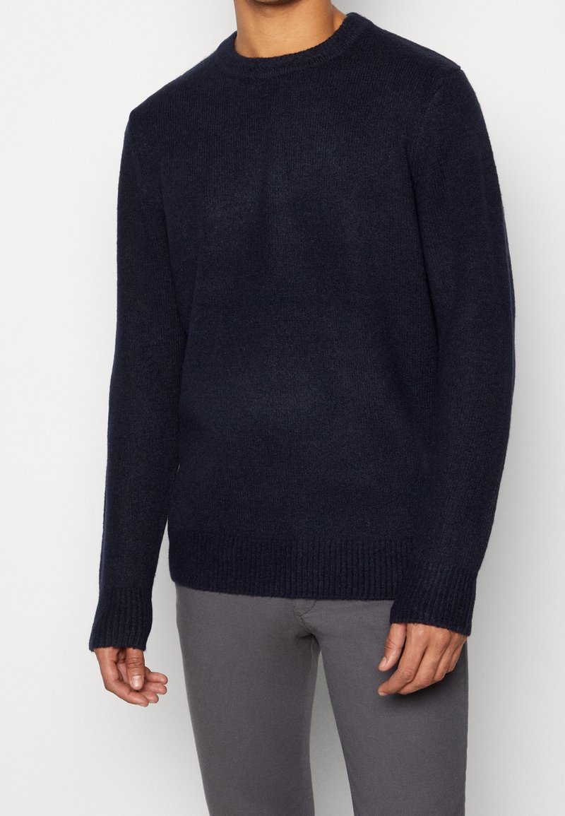 Homme portant un pull en maille bleu marine foncé à manches longues et un pantalon gris, debout devant un fond clair uni.