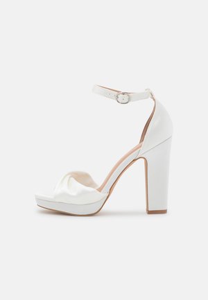 Sandal de salto alto branco com parte superior em cetim e design em nó, faixa no tornozelo com fivela de metal e salto quadrado e robusto.