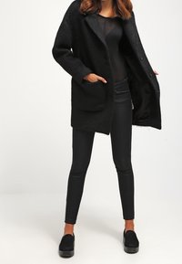 Žena oblečená v černém chlupatém kabátu, průhledném černém topu, černých skinny kalhotách a černých slip-on bot ách, stojící se založenýma rukama v kapsách.