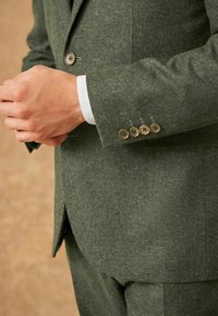 Veste de costume verte en feutre avec une finition texturée, dotée de boutons marron aux poignets et d'un design ajusté.