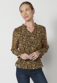 Blusa nera floreale con scollo arricciato. Presenta fiori arancioni e gialli, vita elastica e maniche lunghe arricciate. Abbinata a jeans blu.