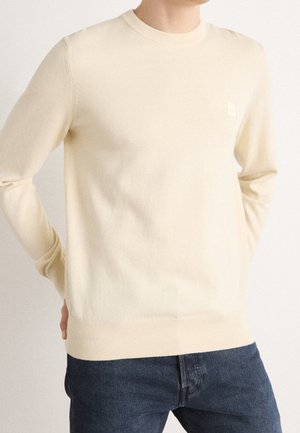 Person trägt einen cremefarbenen Pullover mit langen Ärmeln und dunkelblaue Jeans, steht mit Händen hinter dem Rücken vor einem einfarbigen Hintergrund.