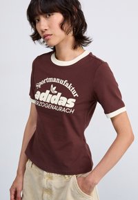 Brown cotton t-shirt with cream-colored collar and sleeve hems; features large white adidas logo and text "Sportmanufaktur Herzogenaurach."