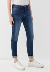 Pantalones vaqueros ajustados de azul oscuro con cintura de talle medio, que presentan un diseño clásico de cinco bolsillos y puños doblados, combinados con zapatillas blancas.