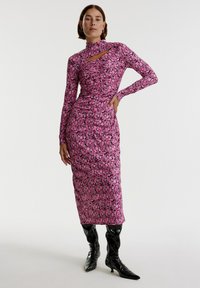 Langärmliges pinkes Blumenkleid mit hohem Kragen und Ausschnittdetails, aus dehnbarem Stoff, kombiniert mit glänzenden schwarzen kniehohen Stiefeln.