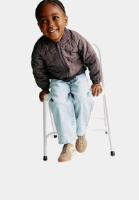 Brun quiltet jakke, lyseblå jeans og tan sneakers. Barn sidder på en hvid stol, smilende, med fletninger.