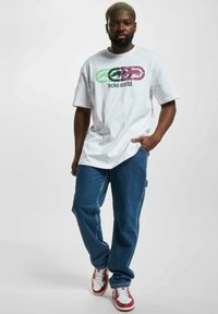 Tricou grafic alb cu logo-uri multicolore, asortat cu blugi albaștri din denim și adidași roșii și albi. Croi casual, cu mâneci scurte.