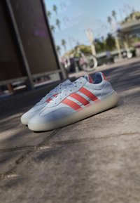 Zapatos deportivos grises y rojos con una parte superior texturizada, tres franjas audaces y una suela de goma translúcida, colocados sobre una superficie pavimentada.