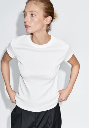 T-shirt bianco in cotone con colletto rotondo e maniche corte, abbinato a pantaloni plissettati neri. Texture liscia, silhouette aderente.