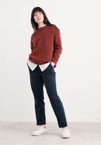 Rūdzīnu krāsas adīta džemperis ar apaļu kaklu, valkāts virs baltas kreklu ar apkakli, kombinēts ar tumši ziliem biksēm un baltām kedām.