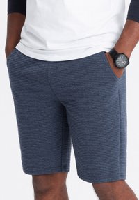Pantaloni corti in tessuto navy con strisce orizzontali, dotati di tasche laterali e vita elasticizzata, abbinati a una camicia bianca e a un orologio nero.
