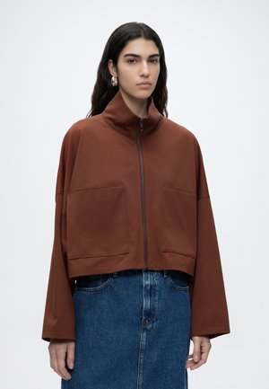 Mujer con cabello largo y oscuro, que lleva pendientes de aro plateados, una chaqueta corta marrón con cremallera y grandes bolsillos delanteros, y una falda vaquera azul.