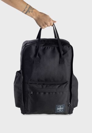 Mochila de nailon negra con dos asas superiores, bolsillo frontal con cremallera, bolsillos laterales y una placa con el logo en la parte delantera. Textura suave y forma rectangular.