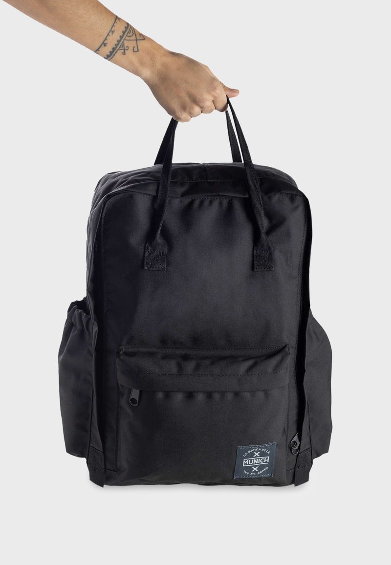 Mochila de nailon negra con dos asas superiores, bolsillo frontal con cremallera, bolsillos laterales y una placa con el logo en la parte delantera. Textura suave y forma rectangular.