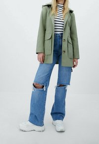 Green lightweight jacket with two front pockets, paired with distressed blue jeans featuring knee rips. White sneakers complete the outfit.