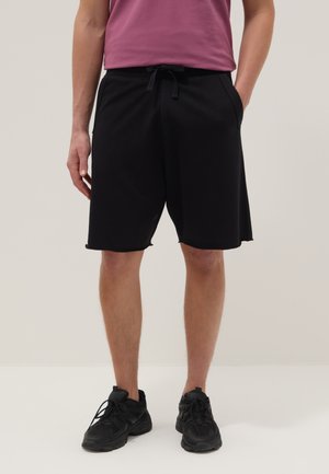Mann trägt schwarze Shorts mit Kordelzug, schwarze Sneakers und ein mauvefarbenes Hemd, steht mit den Händen in den Taschen vor einem schlichten Hintergrund.