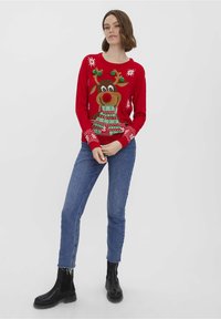 Pull rouge avec un grand motif de renne portant une écharpe verte, des pompons et des motifs de flocons de neige. Associé à un jean bleu et des bottes noires.
