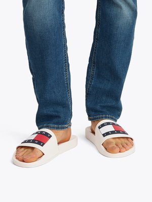 Λευκά πέδιλα με φαρδιά λωρίδα που έχει μαύρο και κόκκινο λογότυπο. Η σόλα είναι επίπεδη και το σχέδιο διαθέτει λείο υλικό. Φορεμένα με μπλε τζιν.