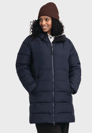 Frau mit lockigem Haar, die eine braune Strickmütze und einen dunkelblauen Steppmantel mit Reißverschluss trägt, die Hände in den Taschen hat, lächelt und zur Seite schaut.