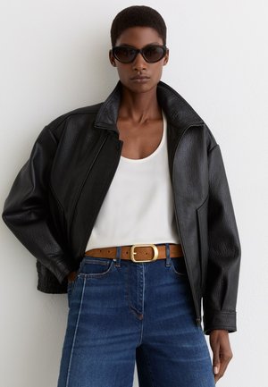 Woman wearing black leather jacket over white top, blue jeans with brown belt, and dark sunglasses, standing against white wall.