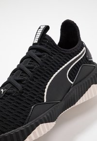 Zwarte gebreide sneaker met een gestructureerde bovenkant, witte accenten aan de zijkant en een dikke, gegroefde zool. Beschikt over een zwarte stoffen tong met branding.