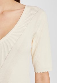 Helles beige, geripptes Strickoberteil mit tiefem V-Ausschnitt, kurzen Ärmeln mit gezacktem Saum und diagonalem Strukturdetail über der Brust.