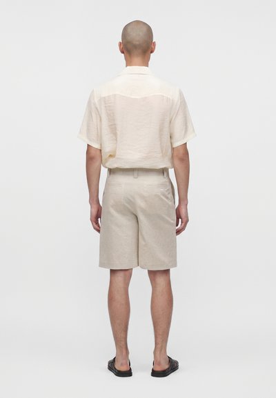 Homme à la tête rasée portant une chemise crème à manches courtes, un short beige et des sandales noires, debout, dos tourné, sur un fond uni.