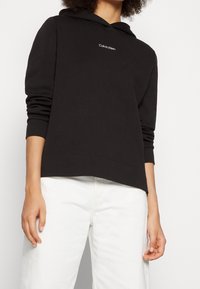 Persona che indossa un hoodie nero di Calvin Klein con logo bianco e pantaloni bianchi, in piedi contro uno sfondo bianco semplice.