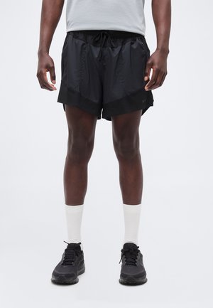 Homme portant un short de sport noir avec un cordon, des chaussettes blanches montantes et des chaussures de sport noires, debout sur un fond uni.