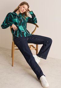 Blusa a maniche lunghe con patttern nero e teal abbinata a jeans scuri e sneakers bianche. Modella seduta su una sedia di legno.