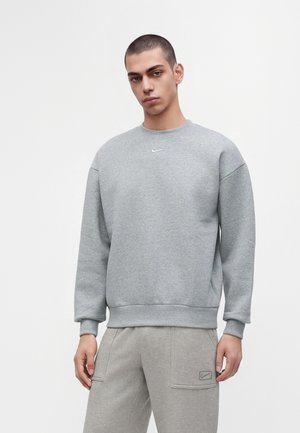 Jeune homme portant un sweat-shirt Nike gris et un pantalon de jogging gris assorti, debout devant un fond clair uni.