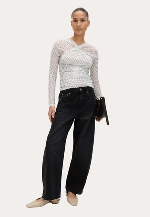 Donna che indossa una maglia bianca trasparente a maniche lunghe con pois, jeans neri a vita alta e gamba larga, ballerine beige, che tiene in mano una pochette nera.