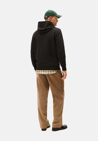 Sudadera negra con capucha y puños acanalados, superpuesta a una camisa de cuadros, combinada con pantalones de pana marrones y zapatos negros. Gorro verde en la cabeza.