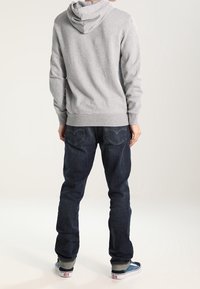 Persona con sudadera gris, jeans azul oscuro y zapatillas azules de pie, de espaldas, frente a un fondo claro y liso.