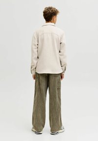 Giovane che sta in piedi di spalle indossando una giacca beige, pantaloni a coste larghi di colore verde oliva e sneakers bianche su uno sfondo neutro.