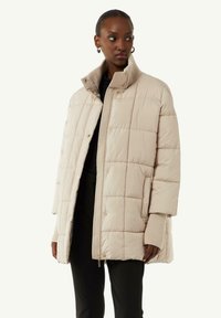 Beżowa pikowana kurtka puchowa z wysokim kołnierzem, przednim zamkiem błyskawicznym i zapięciami na zatrzask. Gładka faktura, luźny krój i ściągacze przy rękawach.