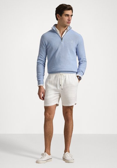Sweater zippé bleu clair avec un tricot texturé et un col ; associé à un short en lin blanc et des baskets en toile beiges.