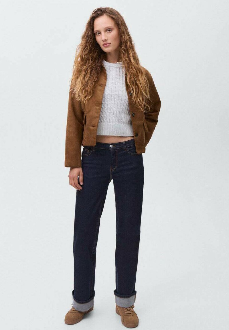 Giacca cropped in suede marrone sopra un maglione grigio chiaro a trecce, abbinata a jeans dritti blu scuro e scarpe beige. Sfondo chiaro.